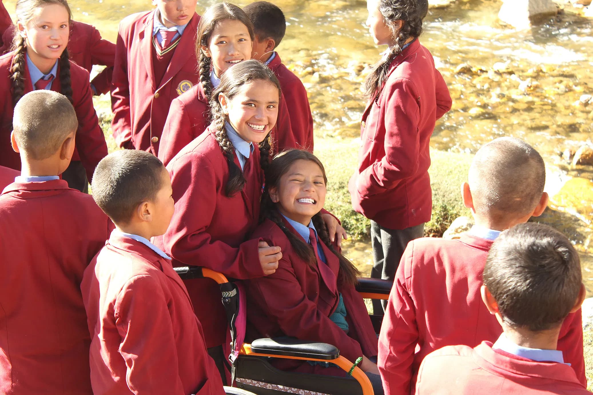 Chuskit in her wheelchair smiling alongside school friends dressed in maroon blazers by a mountain river