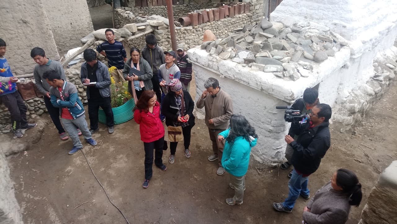Overhead view of director Priya Ramasubban briefing cast and crew near a whitewashed Buddhist chorten in the village