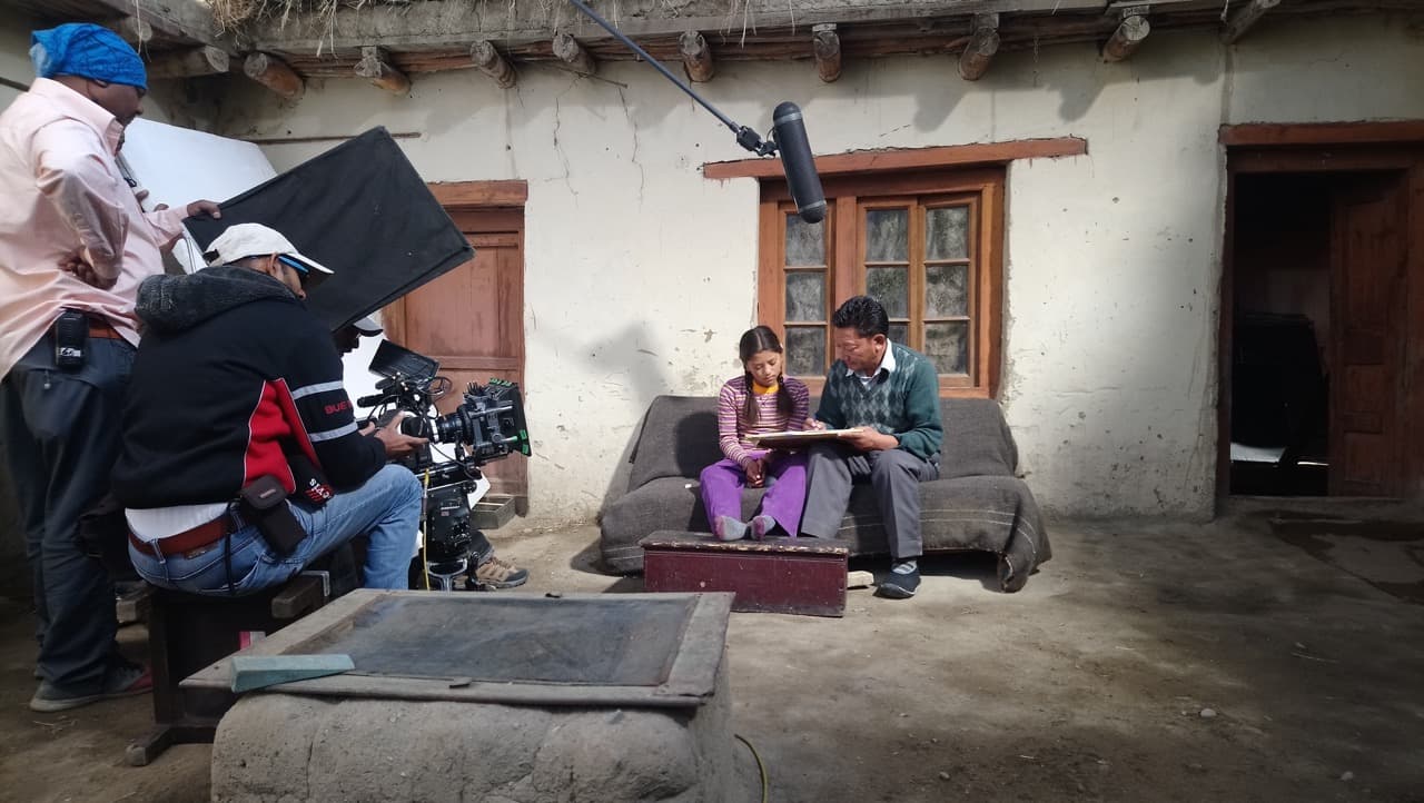 Camera crew and sound recordist capturing a scene of Chuskit and her father studying together on an outdoor courtyard couch