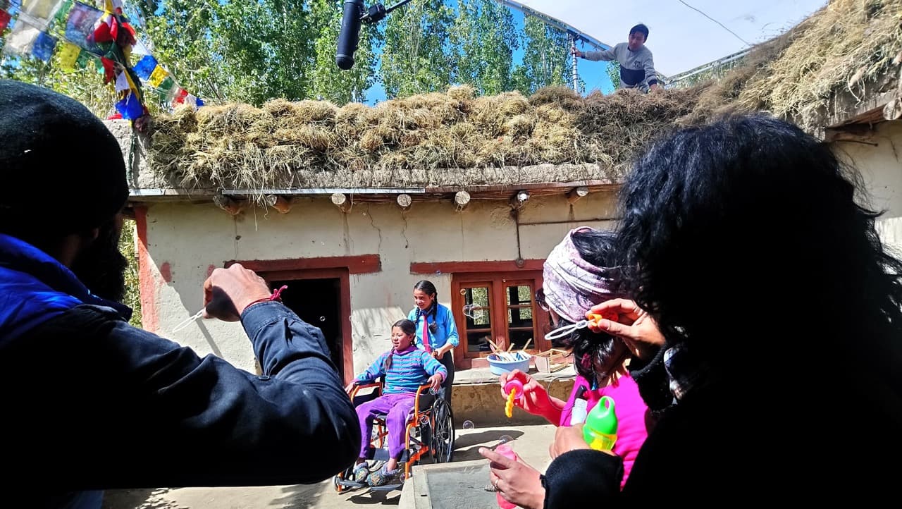 Crew members blowing soap bubbles for Chuskit in her wheelchair while filming in the village courtyard with a rooftop boom mic