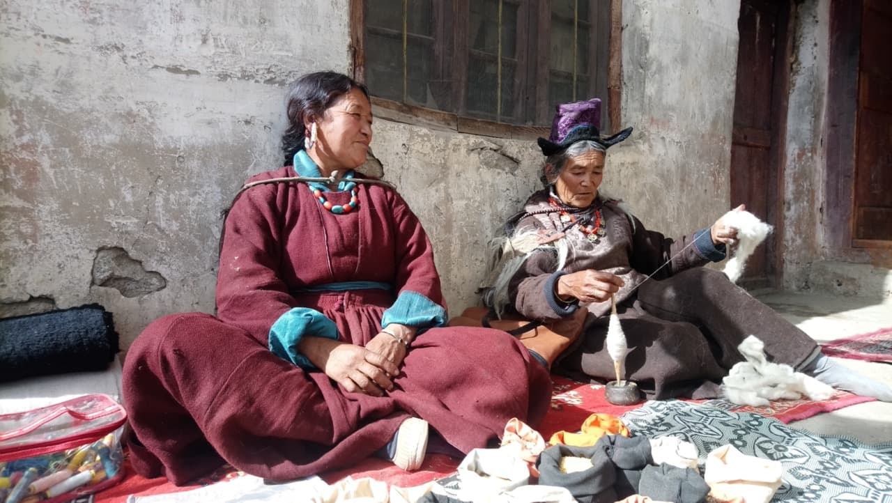Two Ladakhi women in traditional goncha robes and headwear sitting outside spinning wool on a sunny afternoon