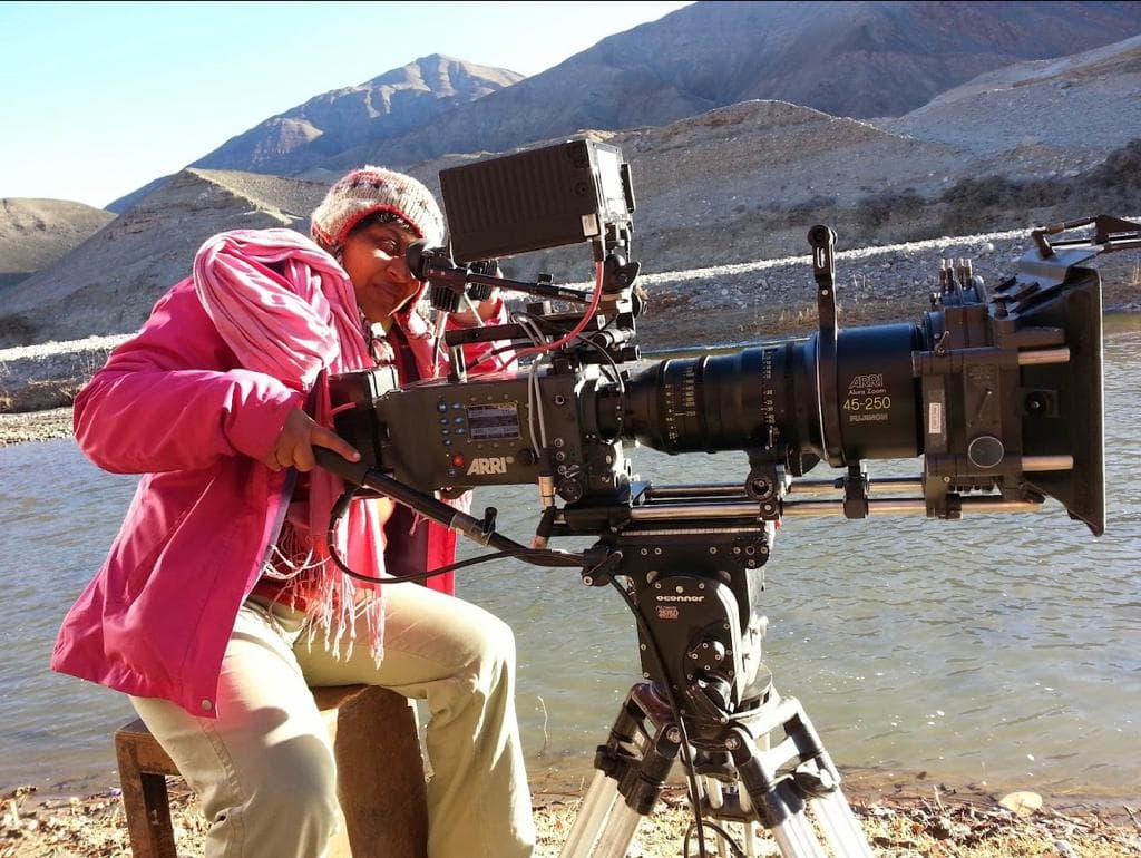 Camera operator peering through the viewfinder of an ARRI cinema camera fitted with an Alura zoom lens beside a river