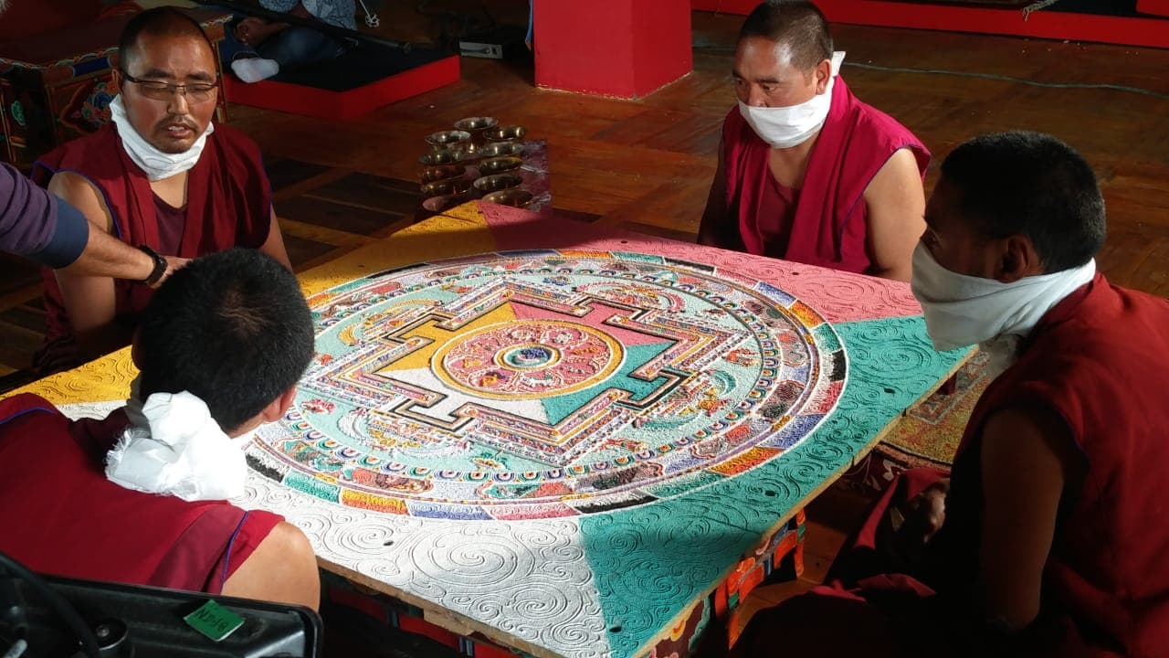 Tibetan Buddhist monks in maroon robes meticulously crafting an intricate sand mandala inside a monastery