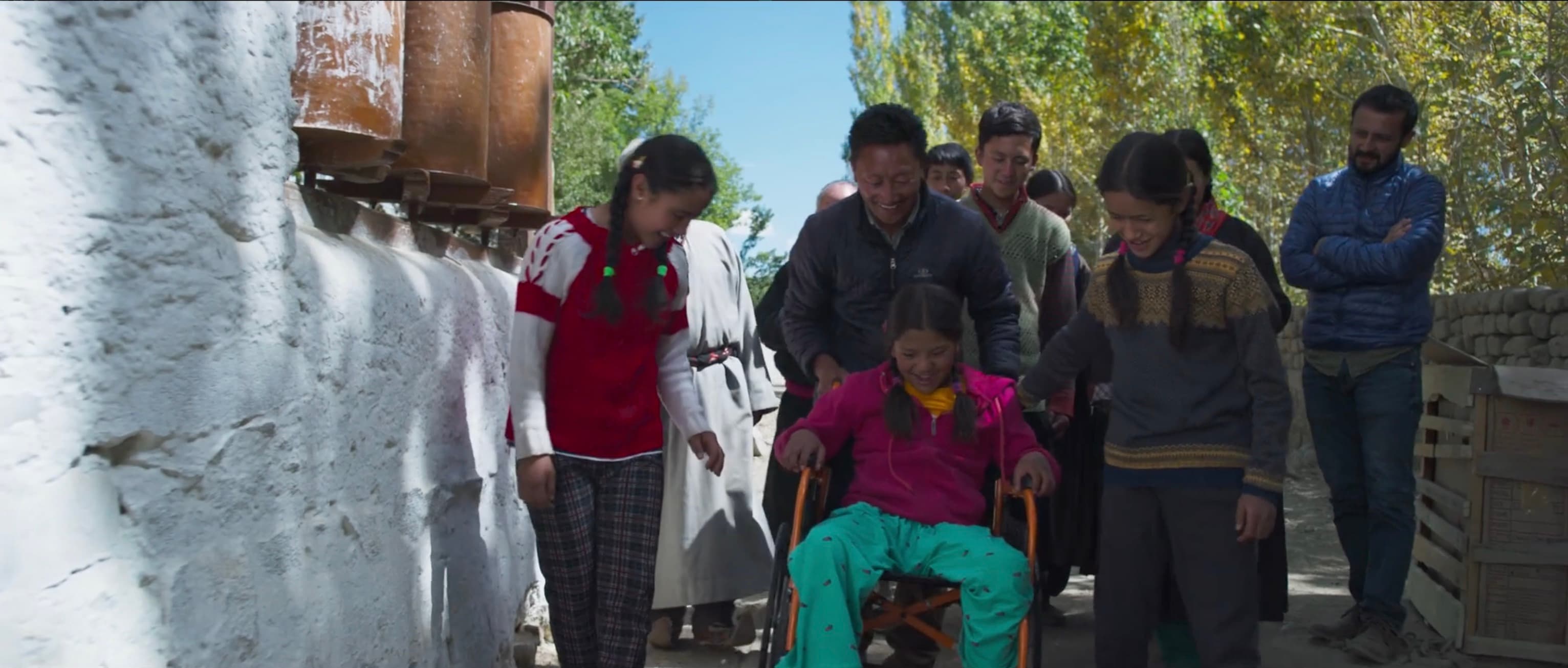 Chuskit being wheeled down a village path past prayer wheels by her father and friends